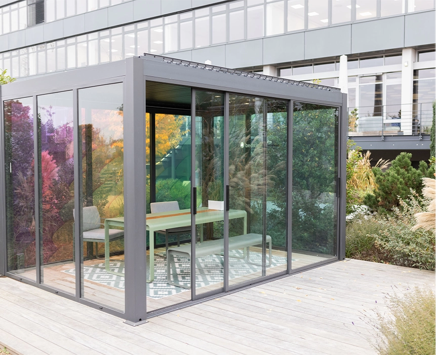 A glass-walled box with sliding doors stands in the middle of a green space; inside, there is a table, a bench and two chairs, surrounded by plenty of plants.