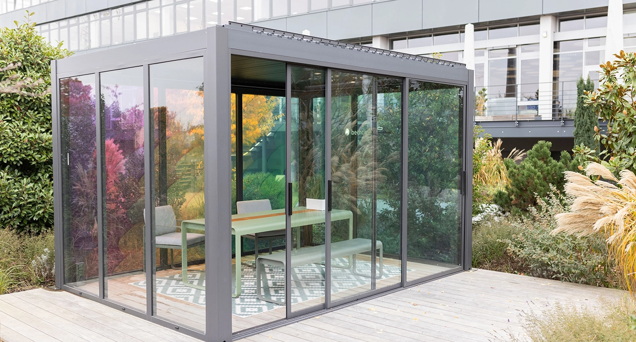 A glass-walled box with sliding doors stands in the middle of a green space; inside, there is a table, a bench and two chairs, surrounded by plenty of plants.