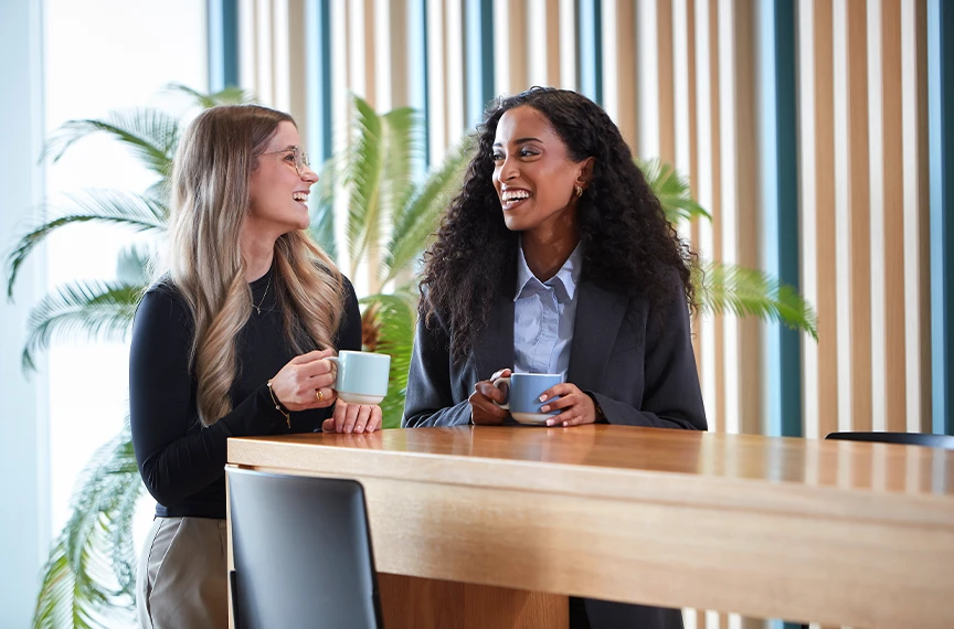 Two women are standing relaxed by a table, holding cups and sharing a laugh.