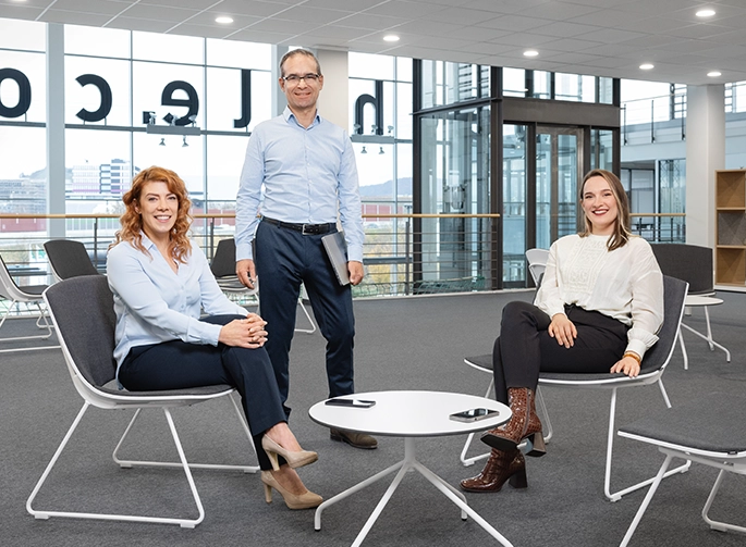 Zwei Bechtle Mitarbeiterinnen sitzen und ein Mitarbeiter steht beim Eingang im Bechtle Büro.