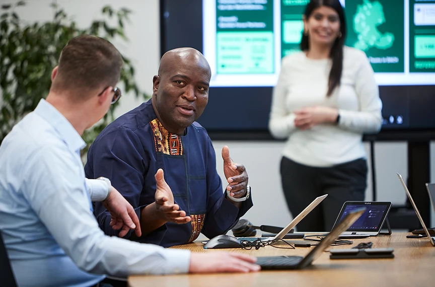 Drei Personen sind zu sehen - zwei führen ein Gespräch vor einem Laptop, während eine dritte Person im Hintergrund vor einem Bildschirm steht.