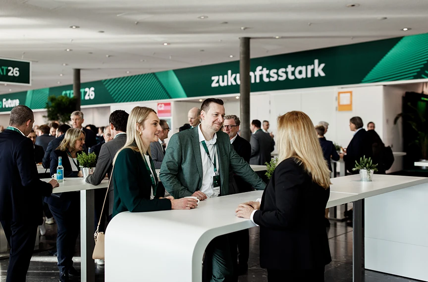 Zwei Frauen und ein Mann stehen zusammen an einem Tisch, wirken aufmerksam und befinden sich vermutlich auf einer Messe.