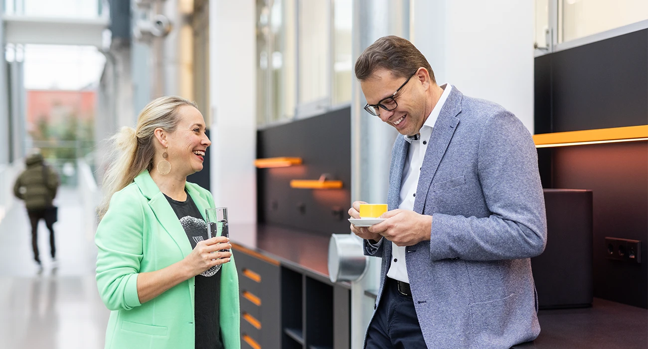 Dies ist ein Foto von zwei Personen in einer professionellen Umgebung, die sich bei einer Tasse Kaffee und einem Glas Wasser unterhalten. 
