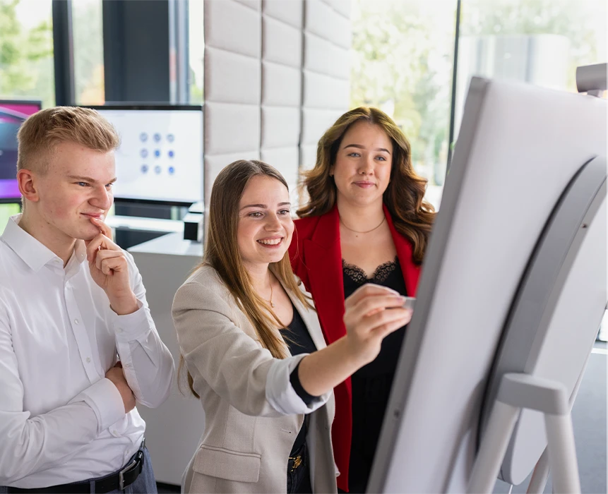 The image shows three smiling individuals in front of a flipchart. The woman in the middle is writing something down. 