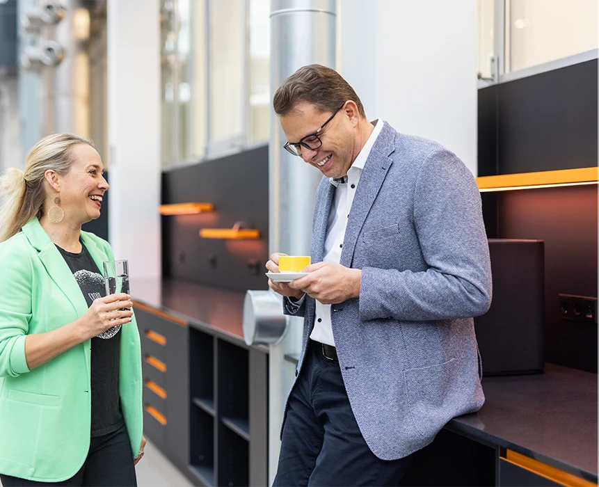This is a photograph of two individuals, in a professional setting, sharing a funny conversation over some coffee and a glass of water. 